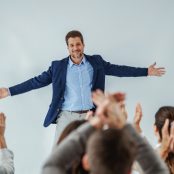 Smiling motivational speaker standing in front of his audience who is clapping.