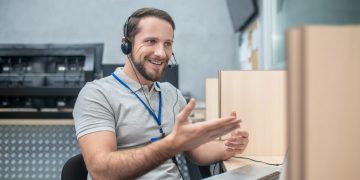 Work, emotion. Young adult smiling gesturing man in headphones near laptop at work place in room